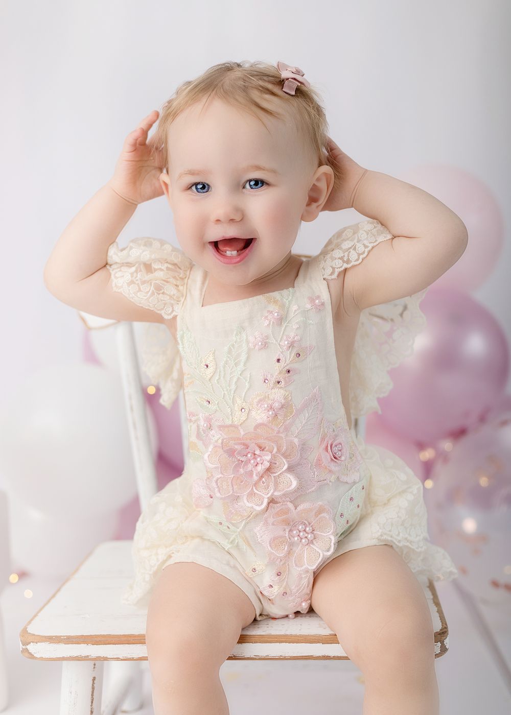 Sweet baby in cream lace romper sitting on chair with pink balloons in dreamy portrait setting.