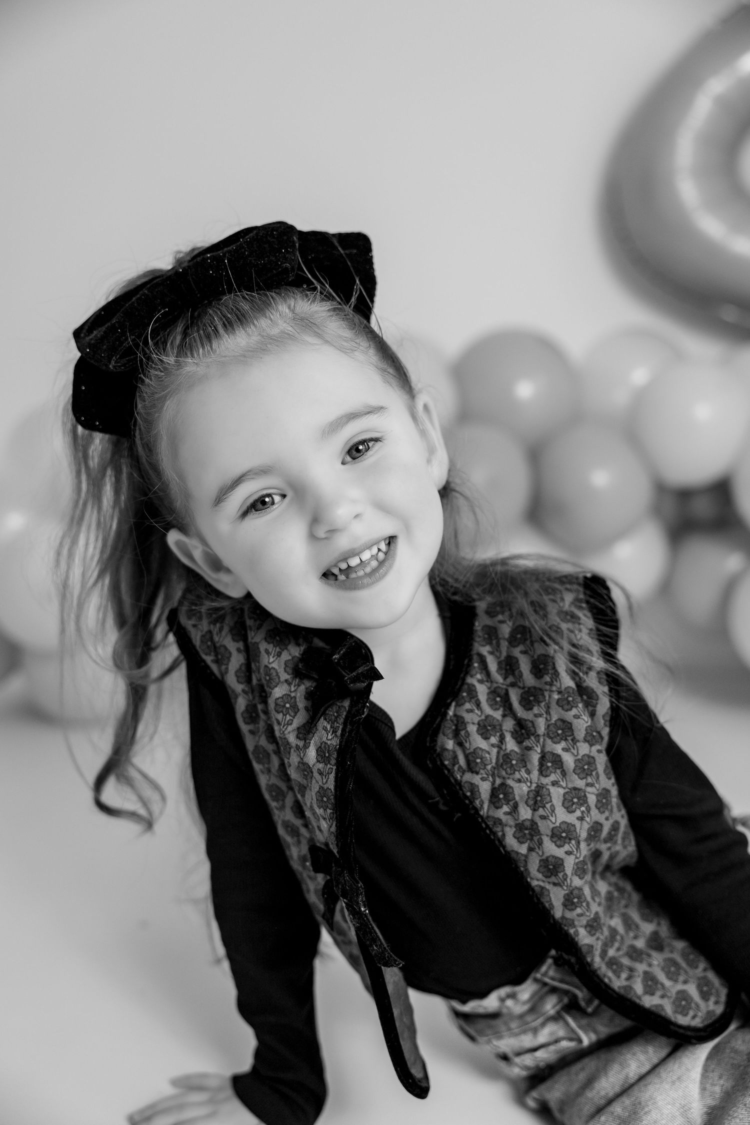 A small baby sits in a classic white clawfoot bathtub surrounded by white balloons in a minimalist black and white photo.