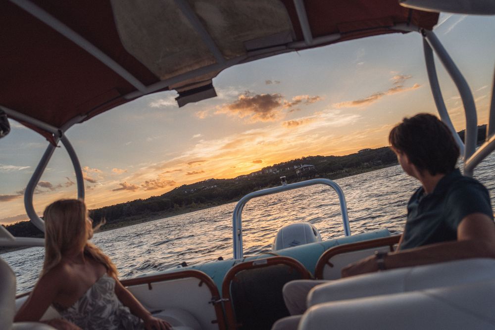 Two people enjoy a scenic sunset boat ride on calm waters with golden skies and silhouetted hills in the background.