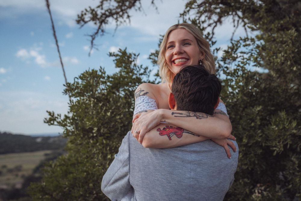 A smiling blonde woman hugs a tattooed man outdoors with lush green trees and blue sky behind them.