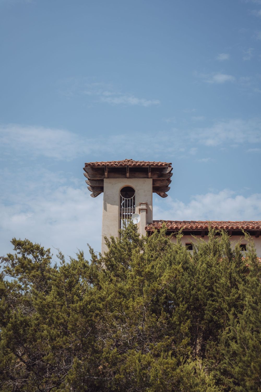 Spanish Colonial Revival tower with red tile roof rises above lush green trees against a blue sky.