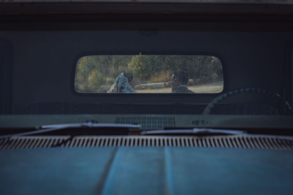 View through a vintage truck's rear window showing two people standing on a rural road surrounded by trees.