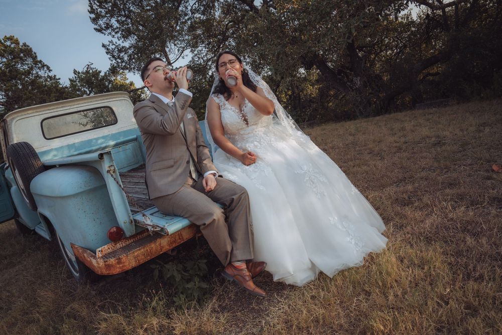 Bride and groom sitting on vintage blue truck tailgate in field, sharing a drink on their wedding day.