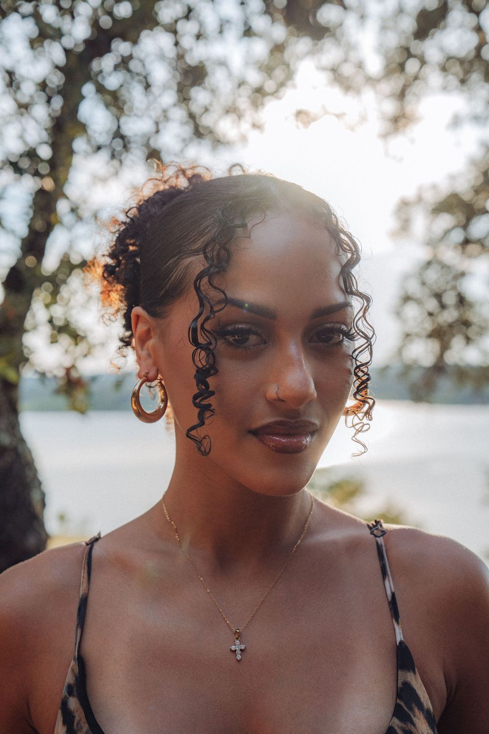 Young woman with curly hair wearing gold hoop earrings and a cross necklace poses outdoors in golden sunlight.