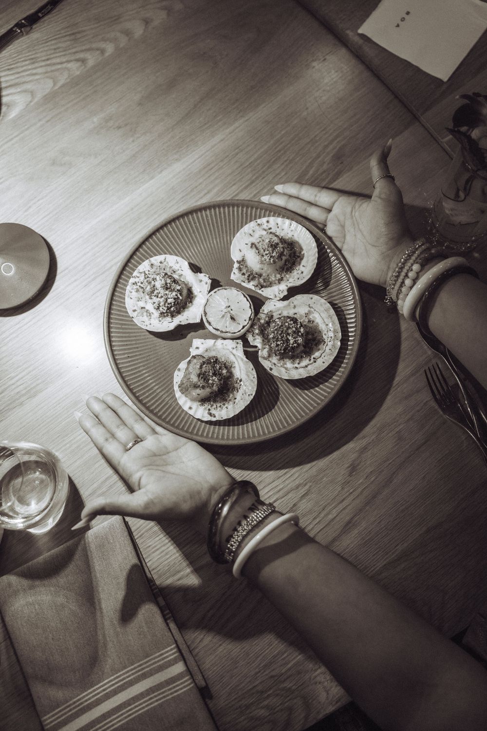 Black and white photo of hands around a plate of stuffed scallops on a wooden table.
