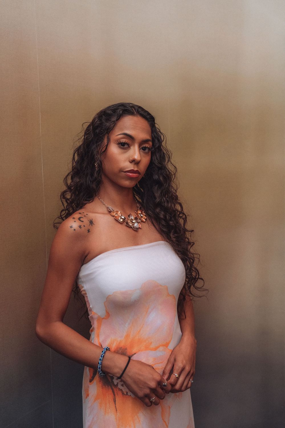 Young woman in white strapless dress with orange print, floral necklace, and curly hair poses against gold wall.