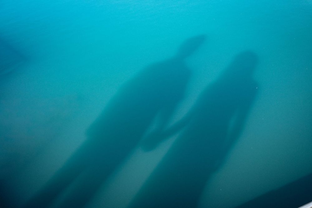 Two shadowy silhouettes of people holding hands reflected on a turquoise underwater surface.