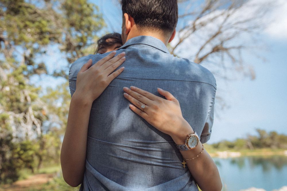 Couple embracing outdoors near a lake, woman wearing a watch hugging man in denim shirt surrounded by trees.