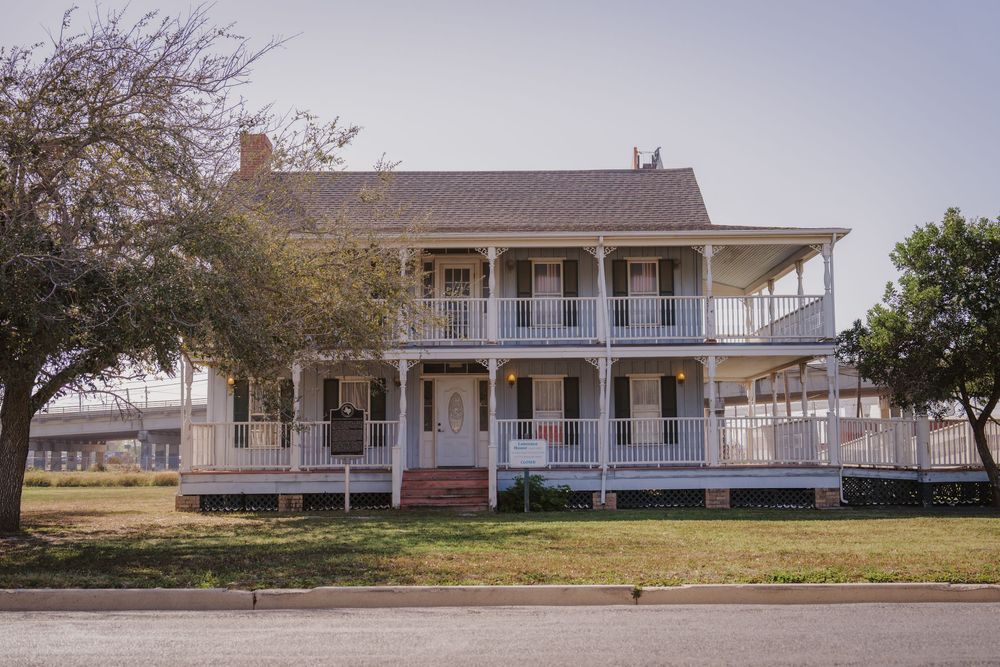 Historic white farmhouse with wraparound porch and brick chimney stands against a rural landscape.