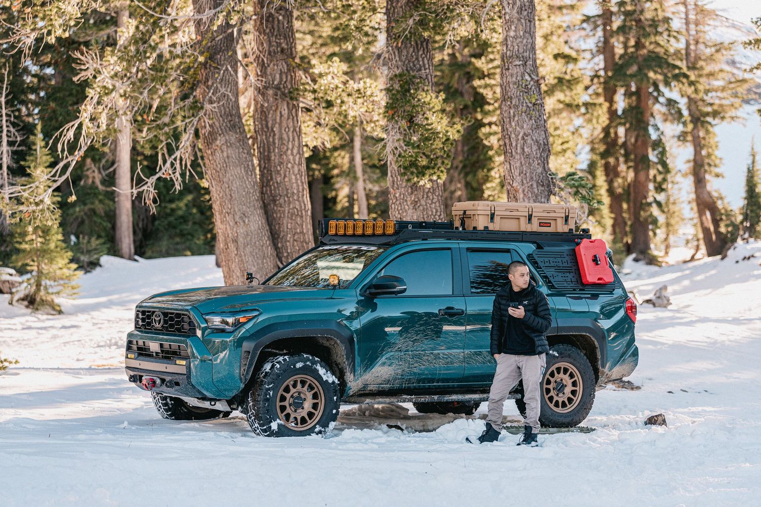 A teal BMW SUV drives through a snowy forest landscape with tall pine trees and winter scenery.