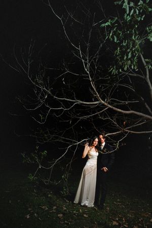 A couple in formal attire stands beneath a dark tree branch at night, illuminated by dramatic lighting.