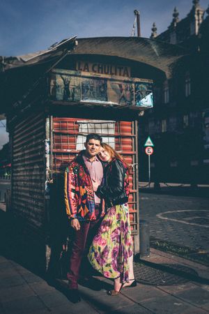 Couple posing in colorful outfits by a vintage newsstand in an urban setting.