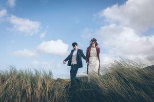 Couple walking through tall grass on hilltop against cloudy sky.