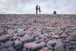 Two figures standing on rocky beach landscape under moody sky.