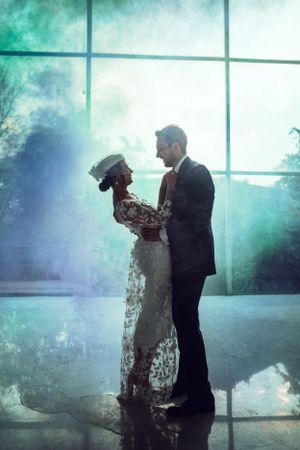 A romantic silhouette of newlyweds embracing in front of large windows with teal smoke effects.