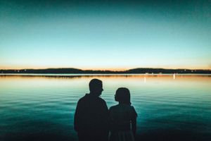 Silhouetted couple sits by calm water at sunset with mountains in the distance.