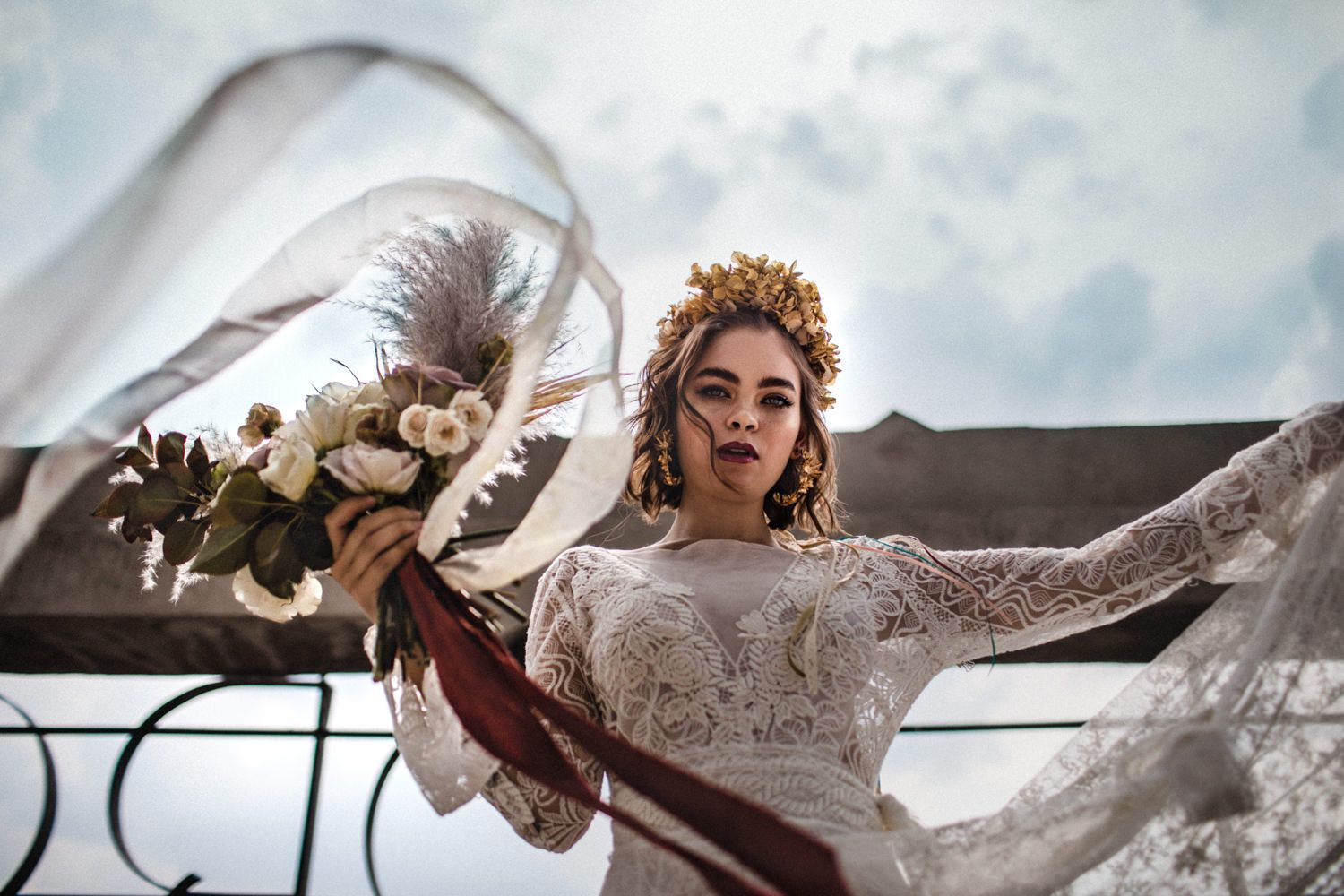 Flowing white wedding dress and veil billow dramatically in the wind against cloudy sky.