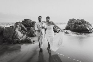 Black and white photo of newlyweds running joyfully along a rocky beach.