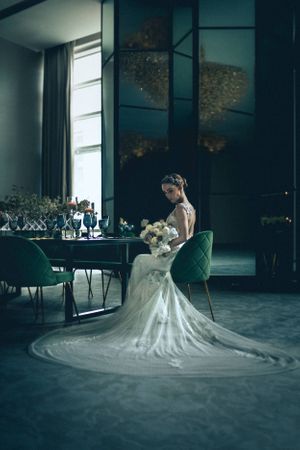 Bride in flowing white gown seated in modern dark room with dramatic lighting.
