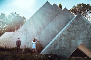 Two people stand against geometric concrete pyramid structures in bright daylight.