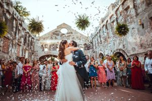 Wedding guests throw confetti as newlyweds walk through stone archways at outdoor celebration.