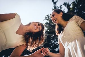Two women in white dresses share a candid laughing moment outdoors.