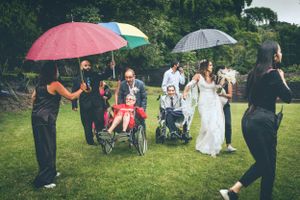 Wedding celebration outdoors with guests holding umbrellas and wheelchair attendees on green lawn.
