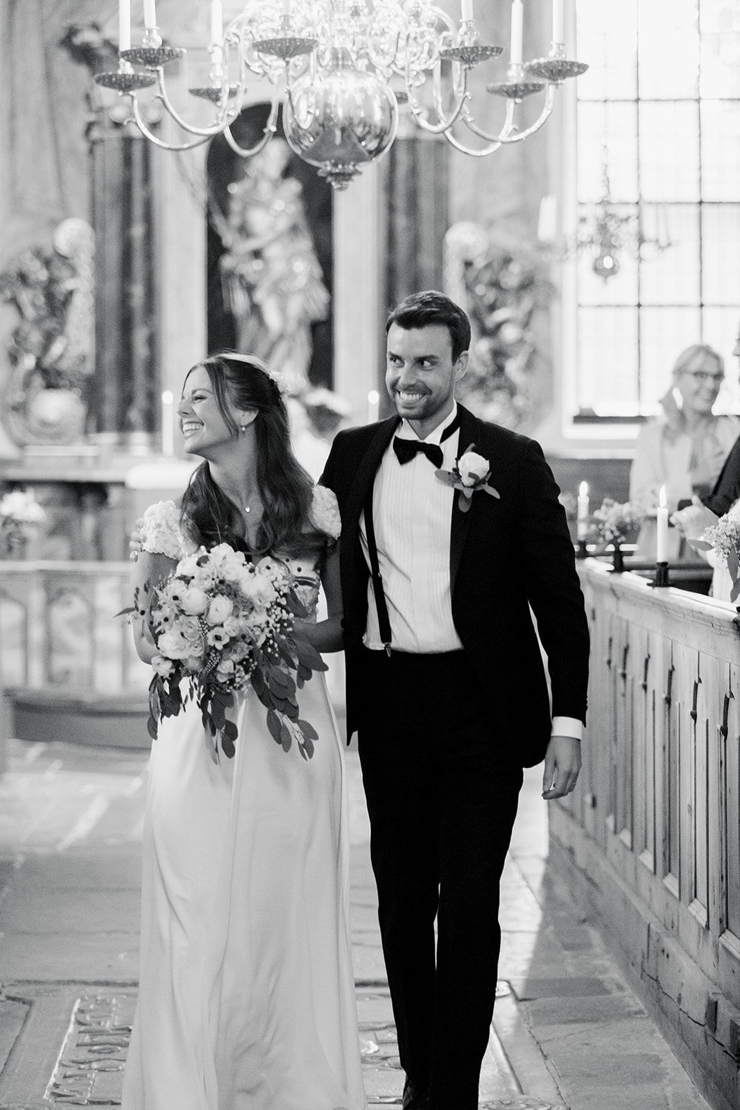 A bride and groom share joyful moments walking down a church aisle with elegant chandeliers overhead.