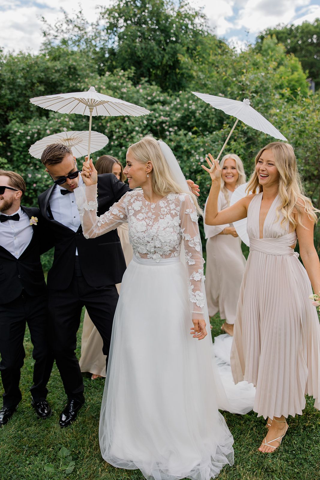 Wedding party in blush pink dresses and black tuxedos pose with white parasols in a lush garden setting.
