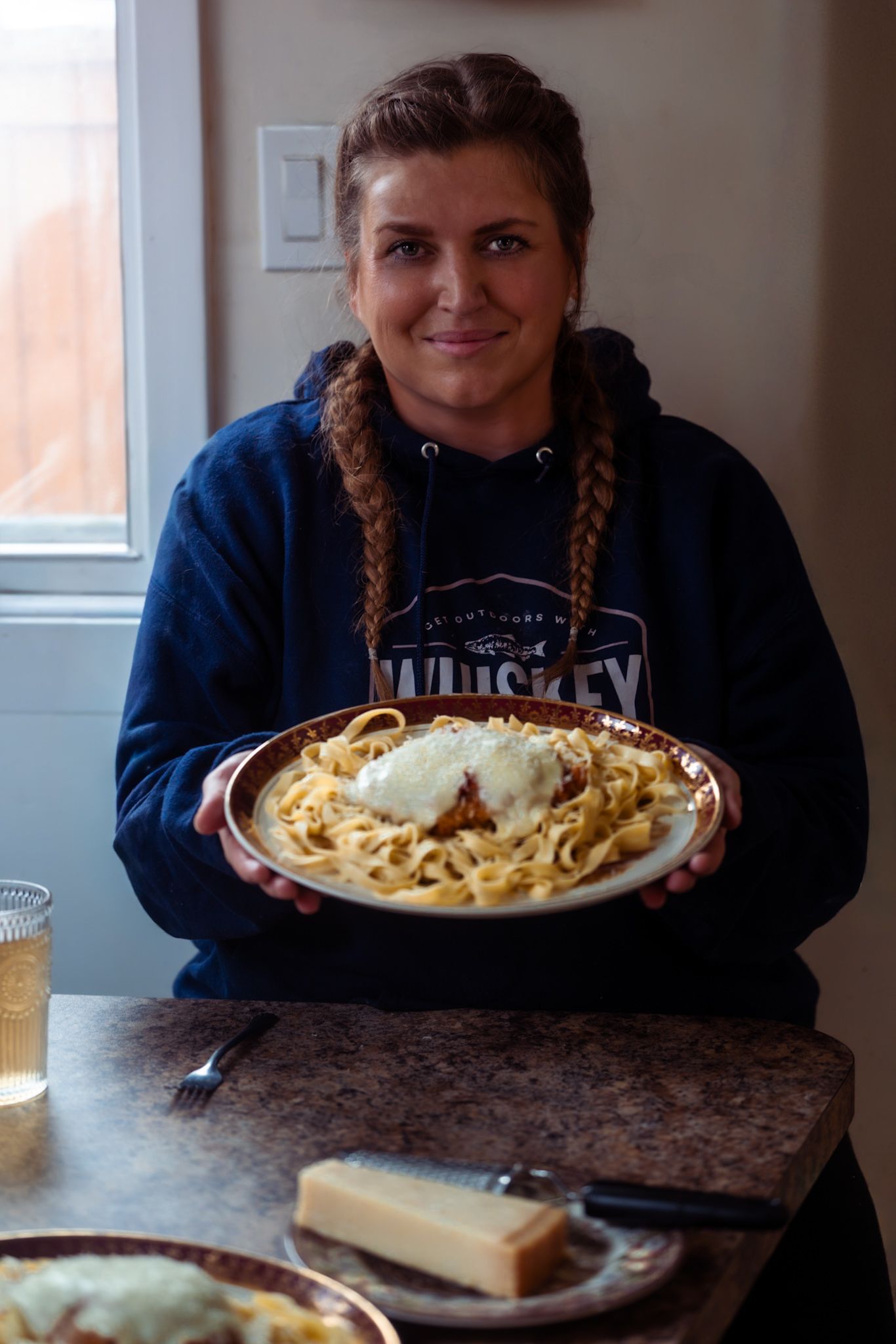 Someone grating fresh parmesan cheese over a plate of fettuccine pasta in a dimly lit restaurant setting.