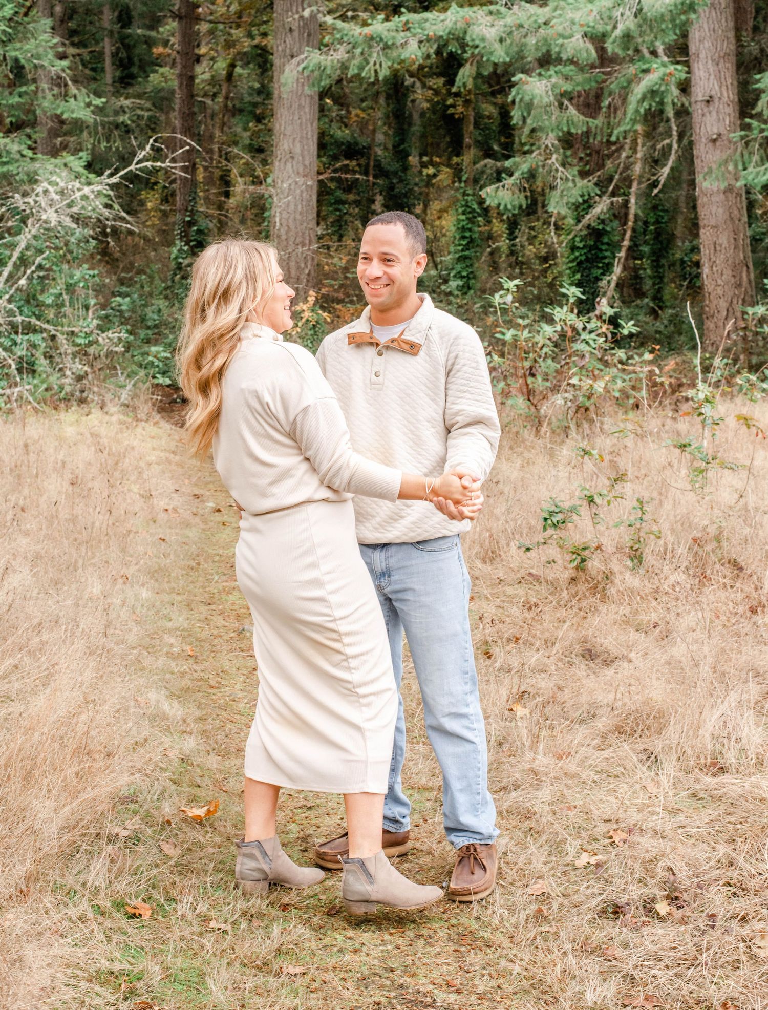 A couple in matching cream sweaters embrace and smile while posing together in a wooded outdoor setting.