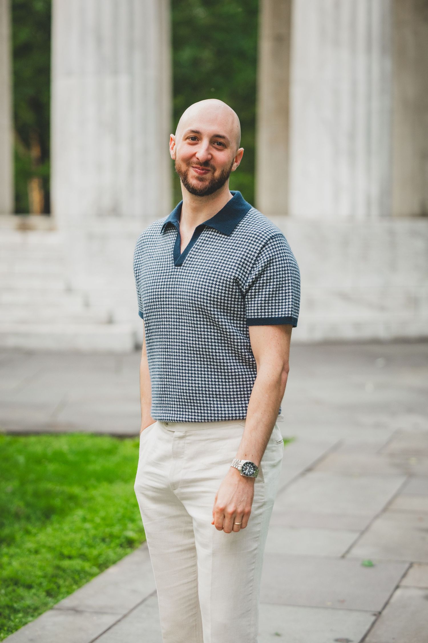 A person wearing a checkered polo shirt and white pants stands near white columns in an outdoor setting.