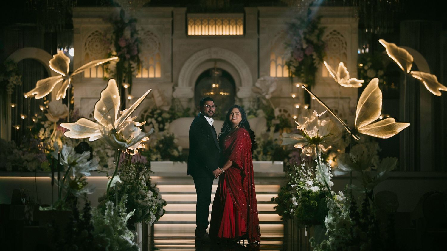 Couple standing together in an elegant hall surrounded by glowing butterfly decorations and lush floral arrangements.