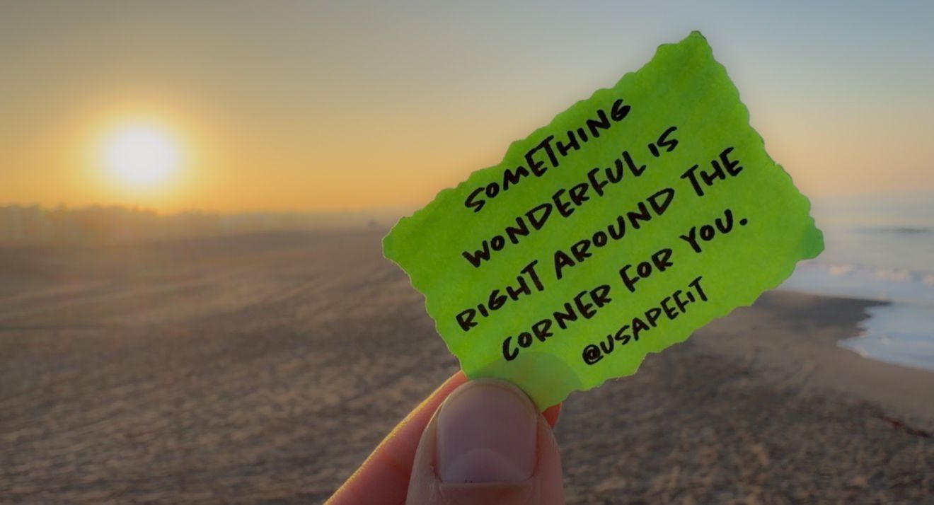 A green sticky note held up against a sunset over an ocean horizon creates a silhouetted view.