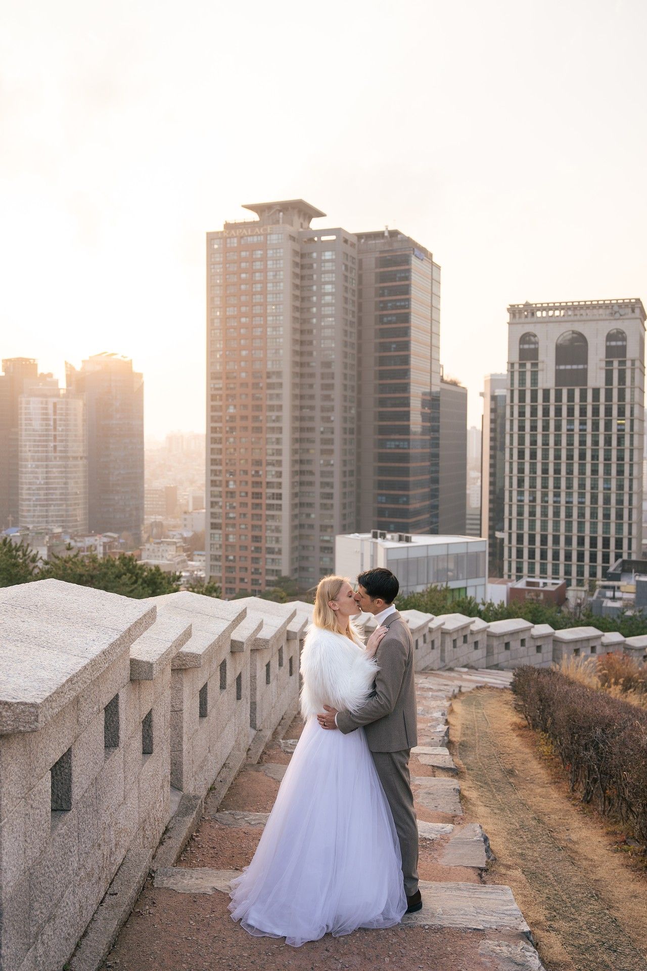 A romantic sunset cityscape view shows a couple in wedding attire walking along an urban wall with high-rise buildings beyond.