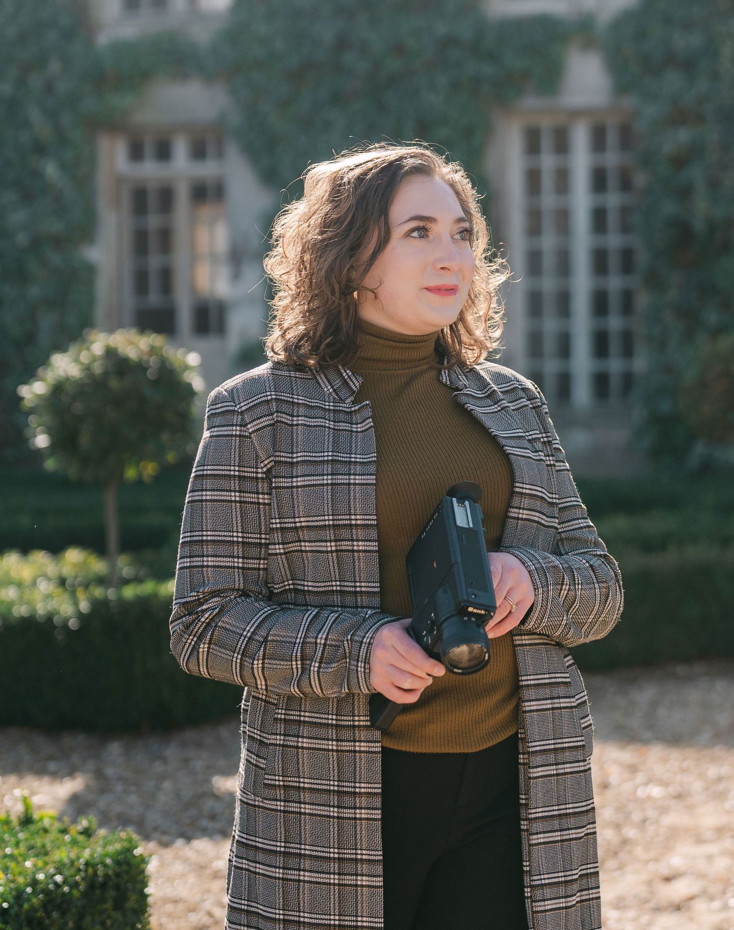 A person wearing a plaid blazer and brown turtleneck sweater poses against a garden backdrop with windows.