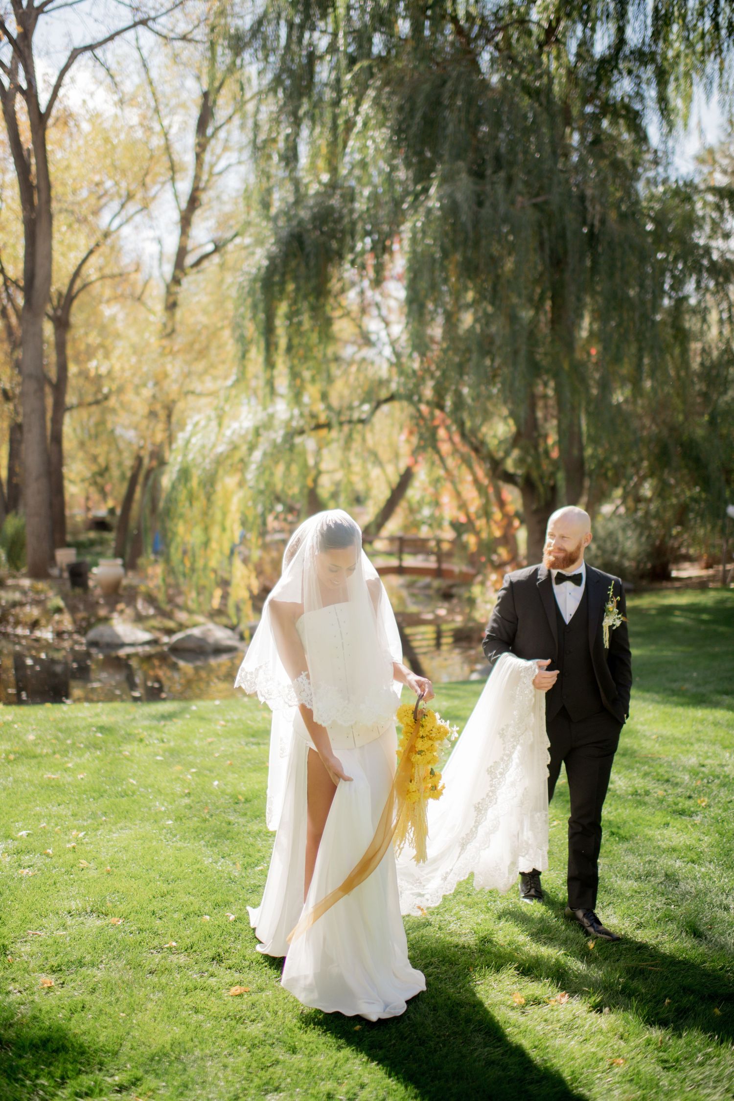 Wedding party walks through a sunlit garden with weeping willow trees casting romantic shadows on the grass.