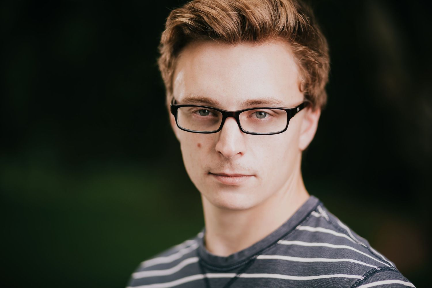 A person wearing glasses and a striped shirt smiles at the camera in an outdoor setting.