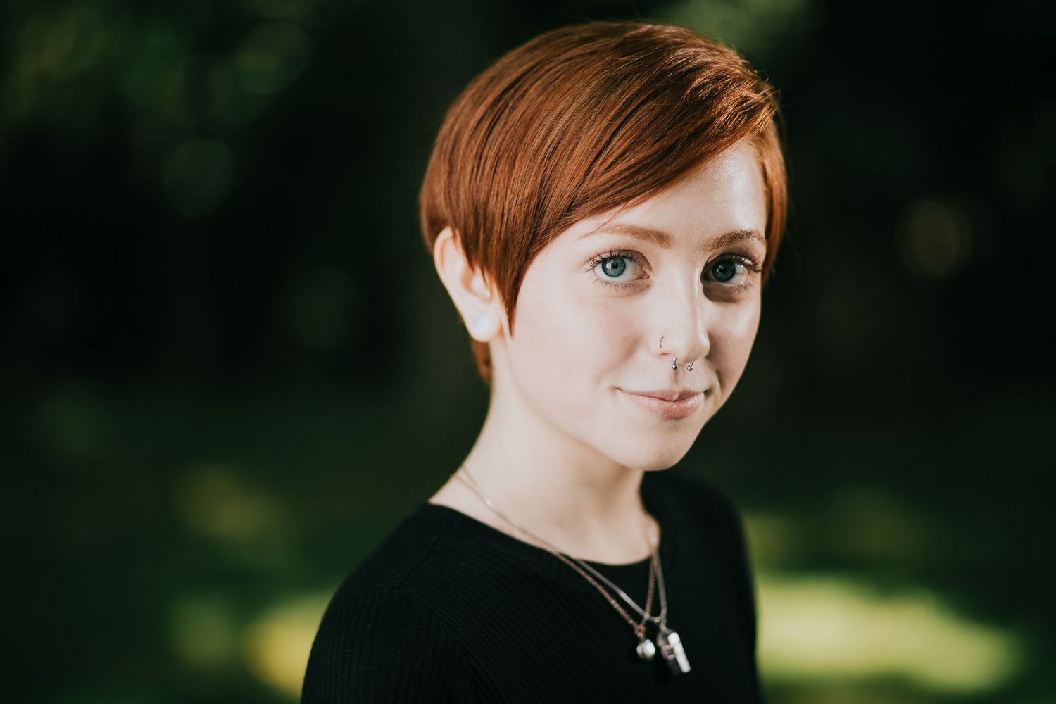 Portrait of someone with short red hair wearing a black top and necklace against a blurred green background.