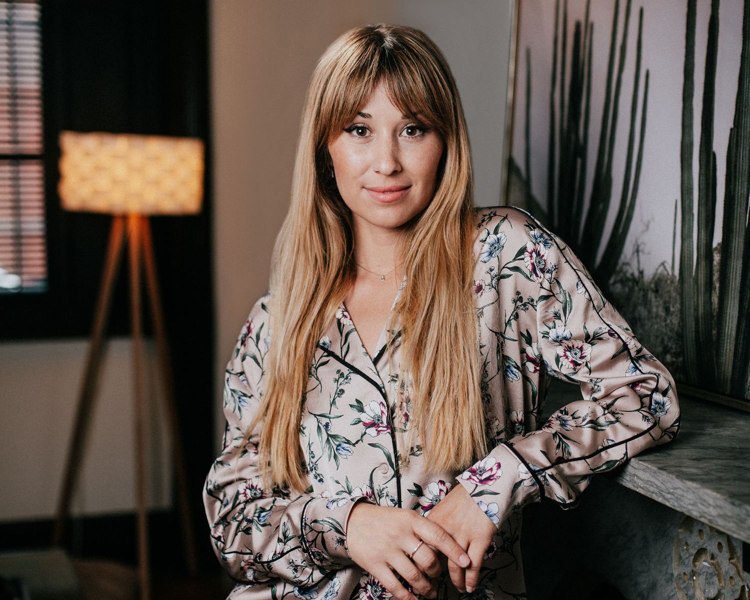 Portrait of someone wearing a floral print blouse with long blonde hair sitting in a room with a floor lamp.