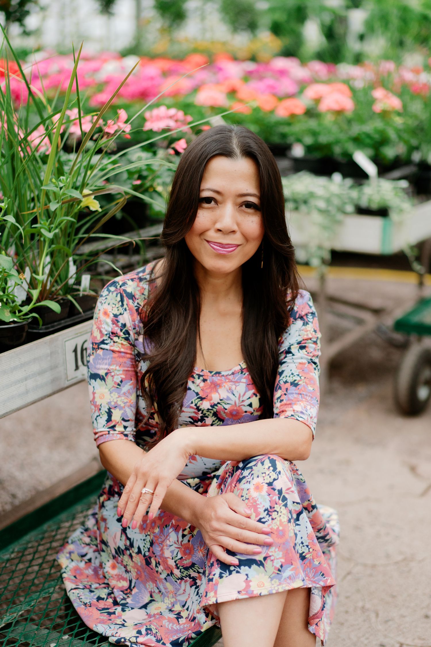 A person wearing a floral pink dress poses in a garden nursery surrounded by colorful flowers and plants.