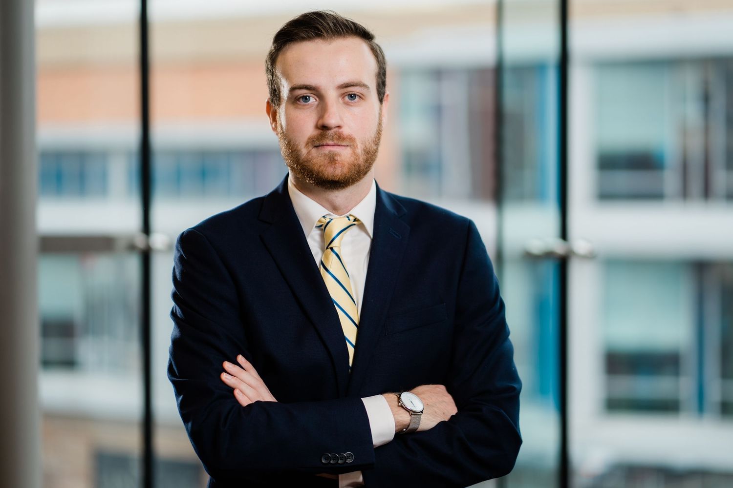 Professional in a dark suit and striped tie stands with arms crossed in front of office windows.