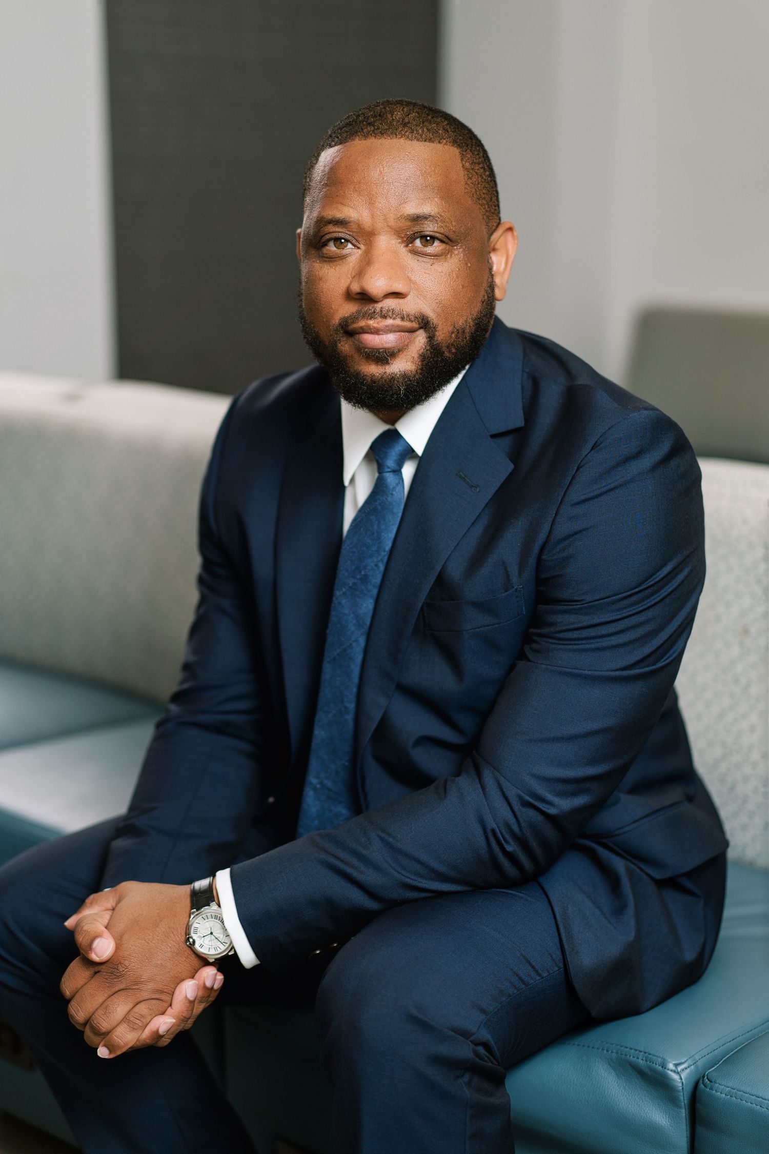 Professional business portrait of person in navy suit sitting on gray couch in modern office setting.