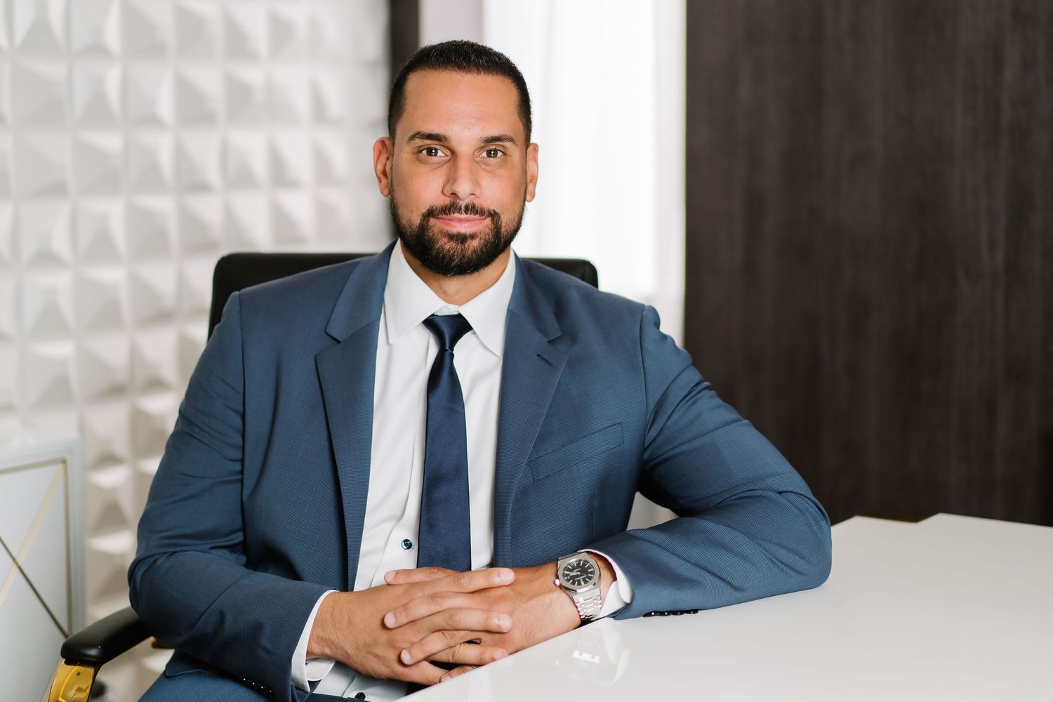 Professional in a navy blue suit and tie sits at desk with hands clasped in modern office setting.
