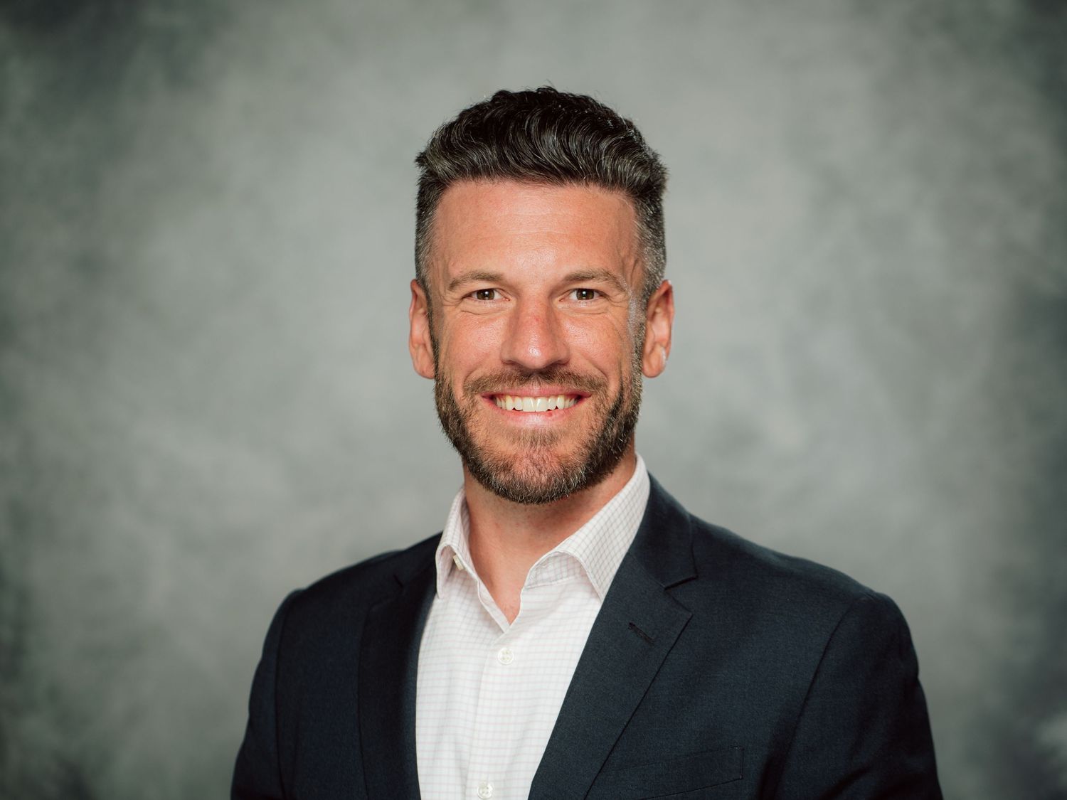 Professional headshot portrait of a person in a dark suit and white shirt against a gray background.