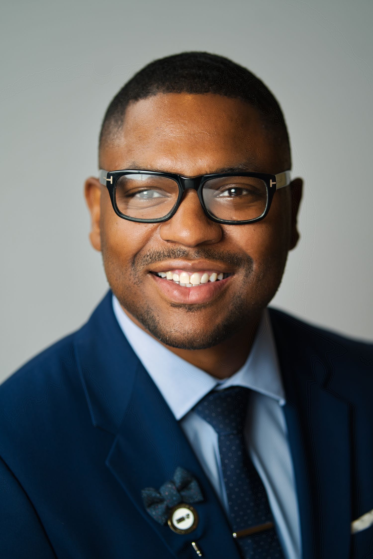 Professional headshot of someone wearing a navy suit, light blue dress shirt, dark tie and black-framed glasses with a warm smile.