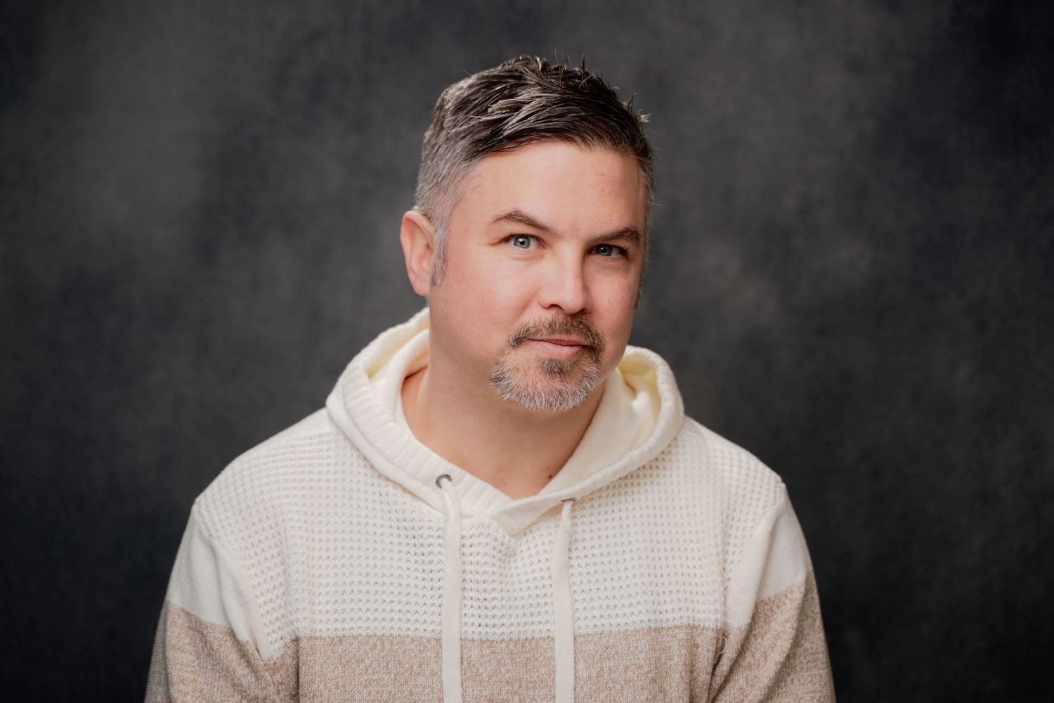 A person wearing a white hoodie poses against a dark background in a professional headshot style photo.