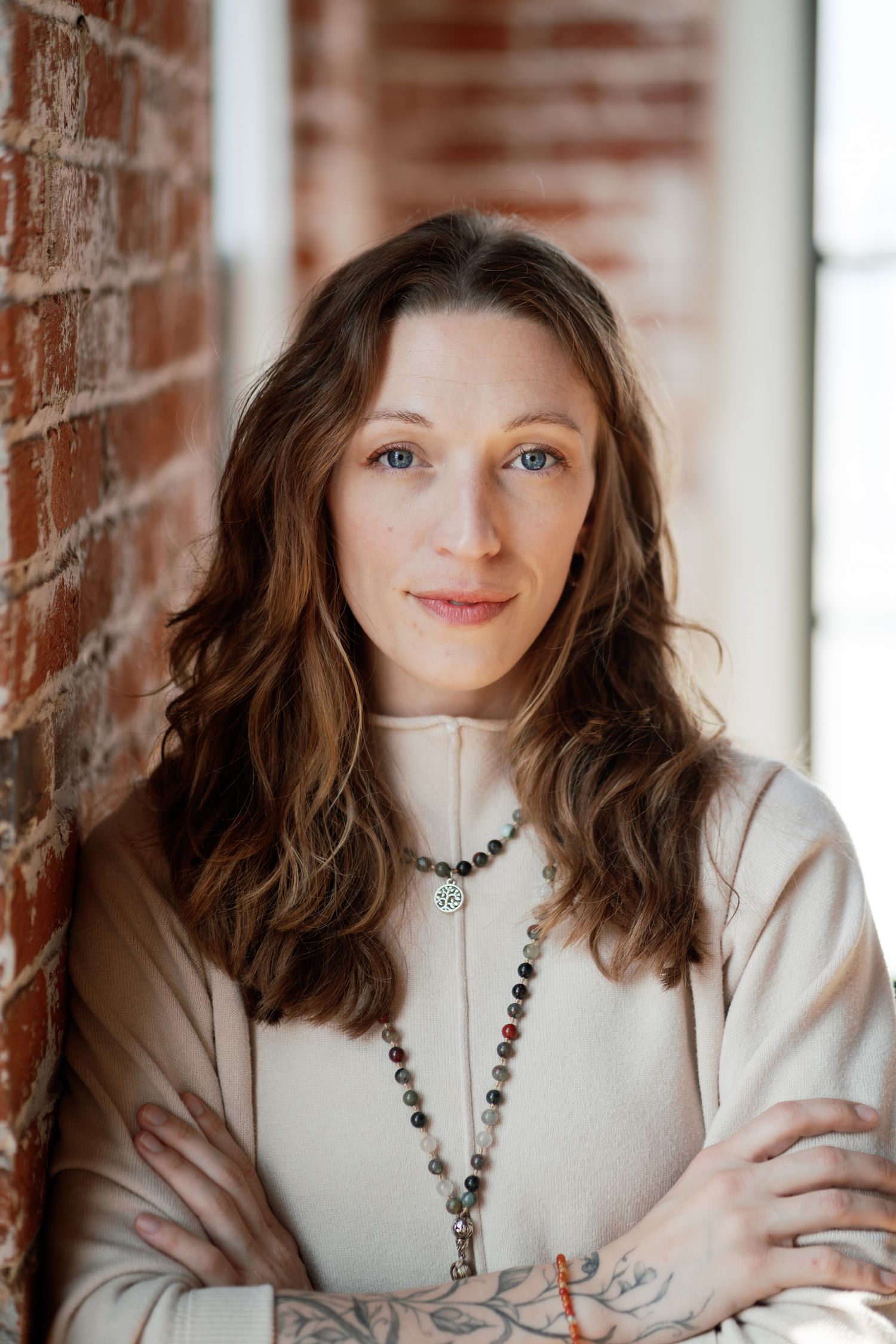 A person in a white top wearing a beaded necklace sits near a brick wall in a bright indoor setting.