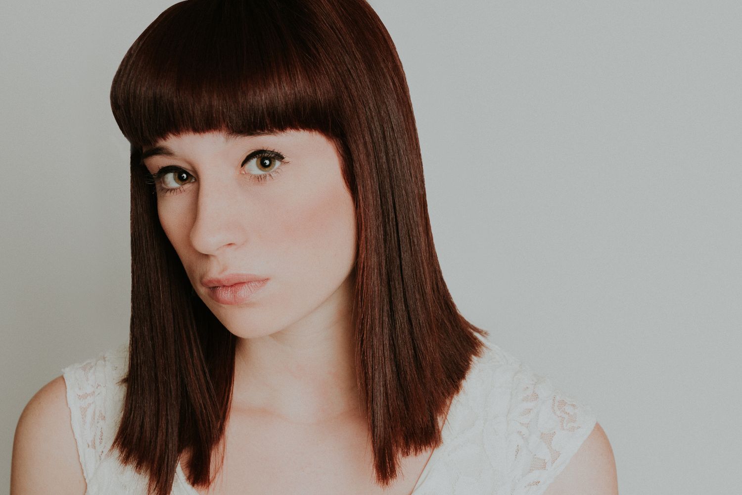 Studio portrait of a woman with straight dark hair and bangs wearing a white top against a light background.