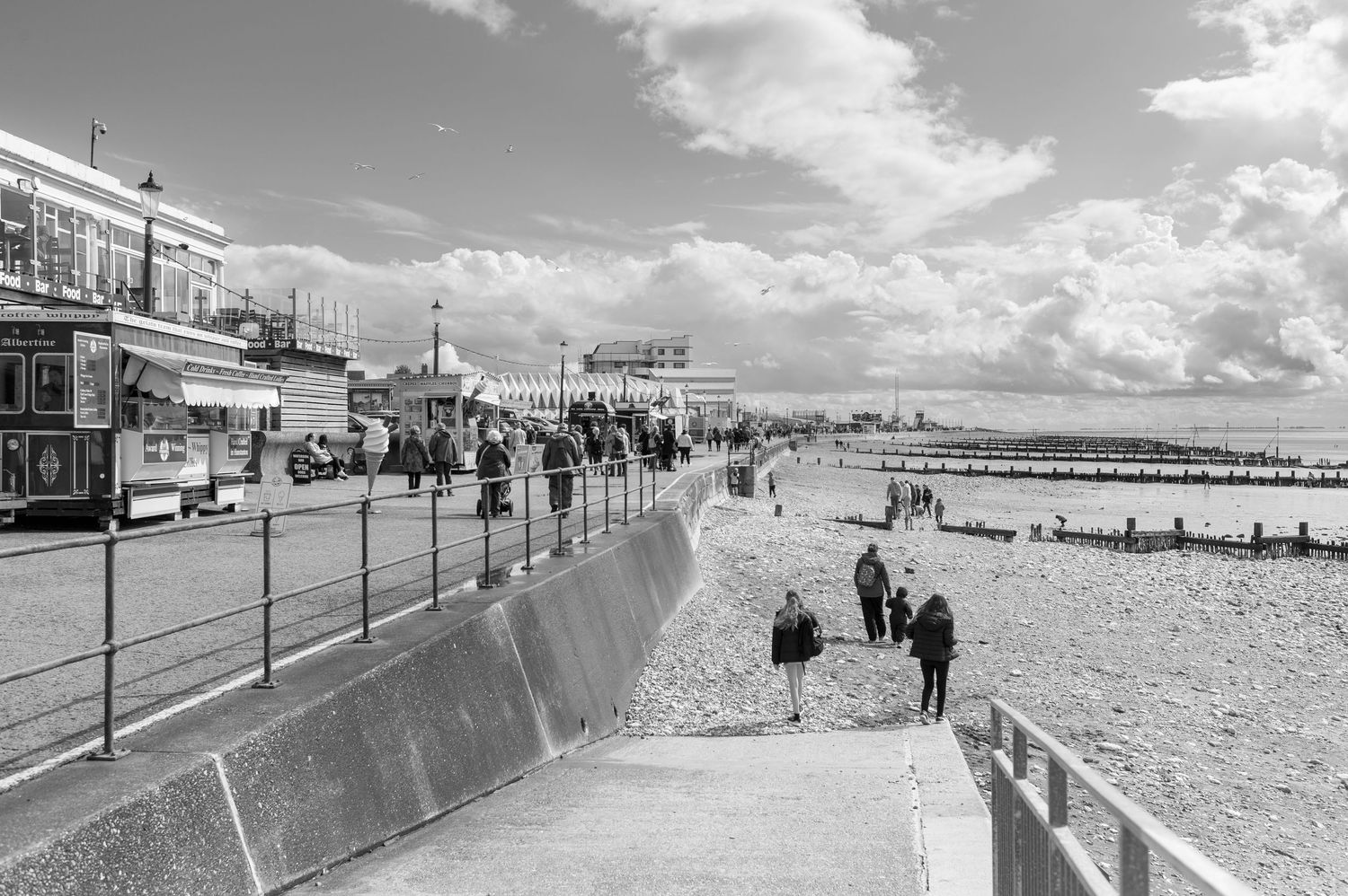 Black and white panoramic view of a beachfront boardwalk with people walking along the promenade on a sunny day.